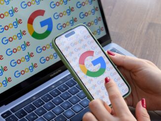 A woman looks at a mobile phone displaying the logo of Google in front of a laptop screen displaying the logo of Google.